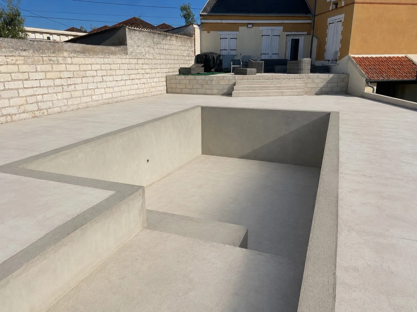 Coque de piscine neuve en béton brut, escalier maçonné, plage béton et terrasse de la maison au fond