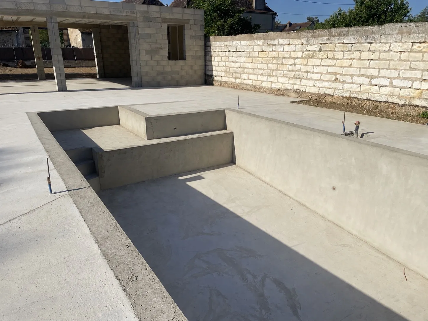 Coque de piscine neuve en béton enduit, escalier maçonné, pool house en parpaings au fond