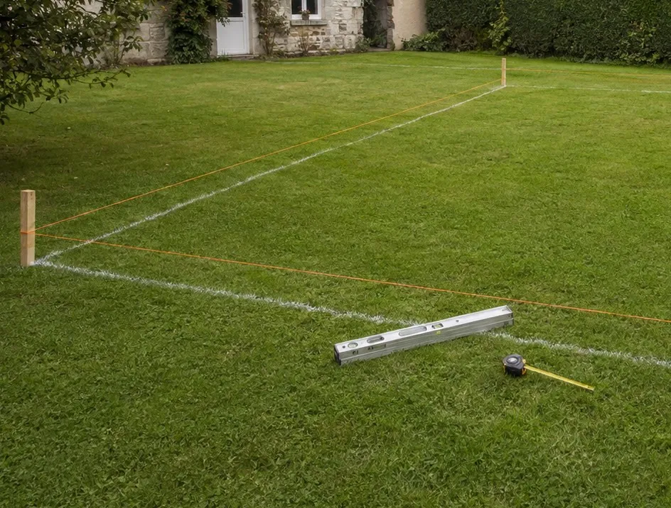 Angle d’implantation sur pelouse : piquet en bois, cordeaux orange tendus, tracé à la chaux, niveau à bulle et mètre posés dans l’herbe